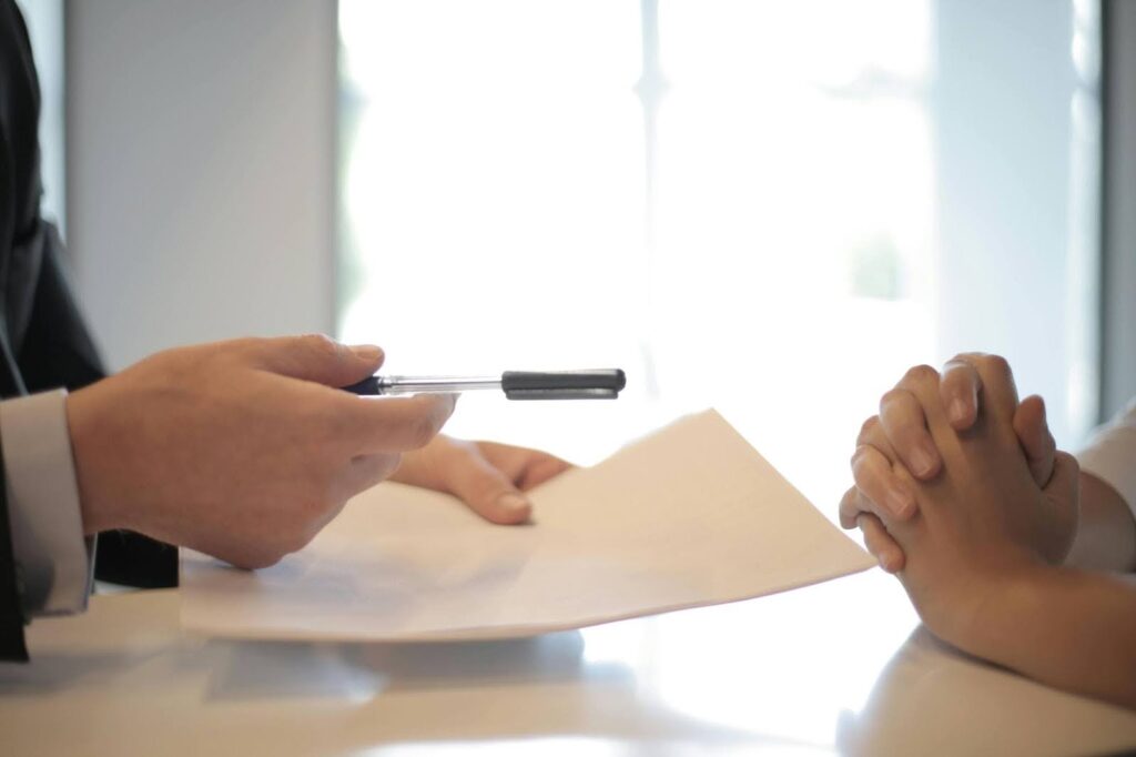 Close-up of a person handing over a pen and contract to another person with clasped hands, preparing to sign a document.
